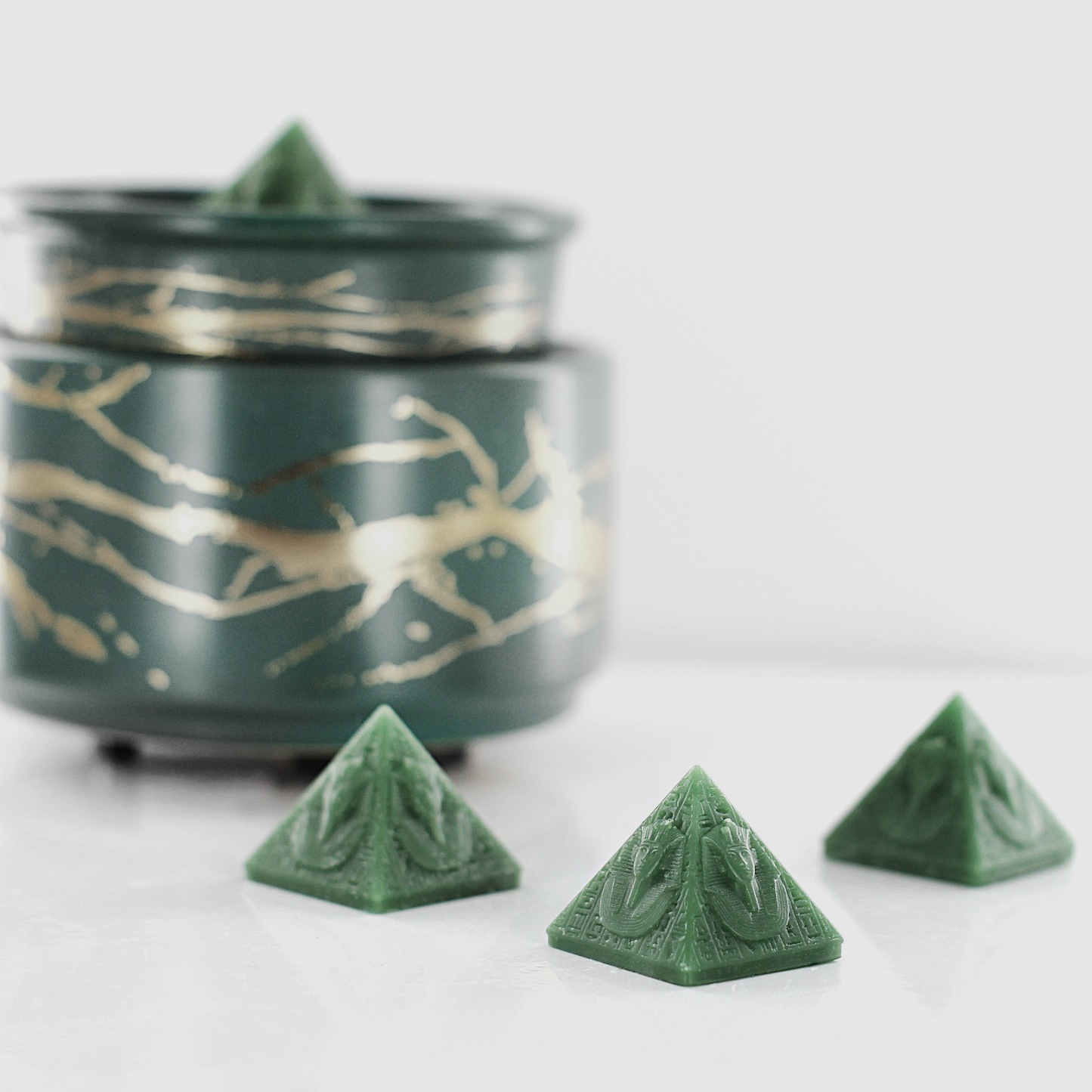 Green fragranced wax melts shaped like pyramids with intricate details, displayed next to a decorative wax warmer with a gold branch design.