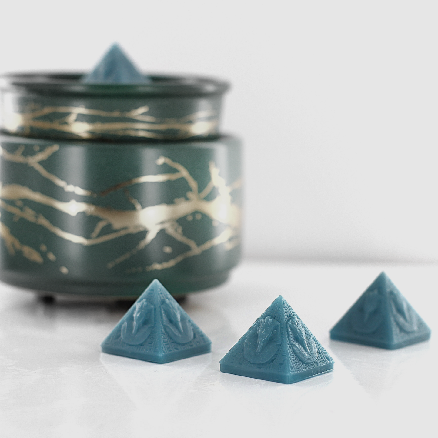 Aqua blue fragranced wax melts shaped like pyramids with intricate details, displayed next to a decorative wax warmer with a gold branch design.