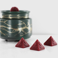 Dark red fragranced wax melts shaped like pyramids with intricate details, displayed next to a decorative wax warmer with a gold branch design.