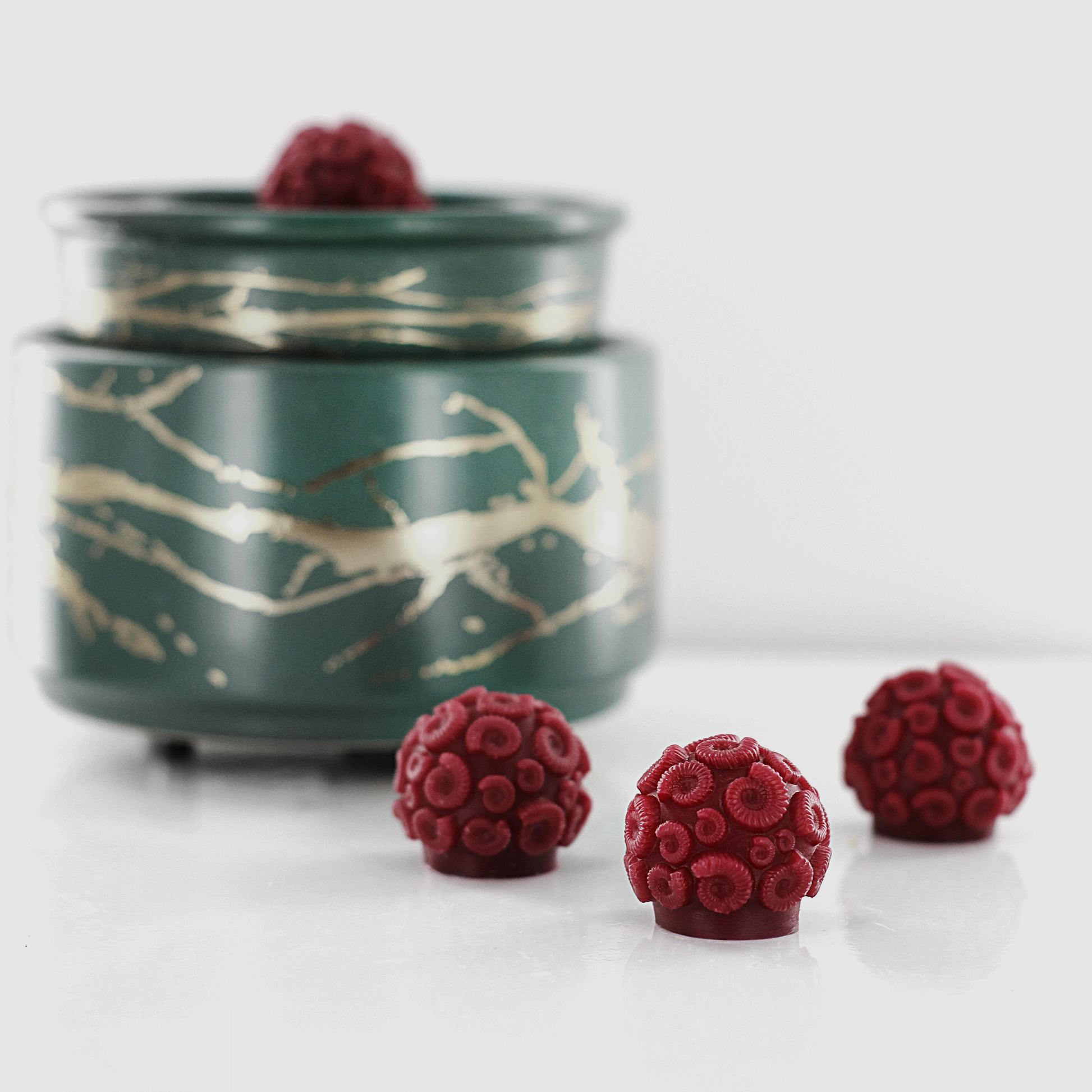 Dark red fragranced wax melts shaped like ammonites with intricate spiral details, displayed alongside a decorative wax warmer with a gold branch design.