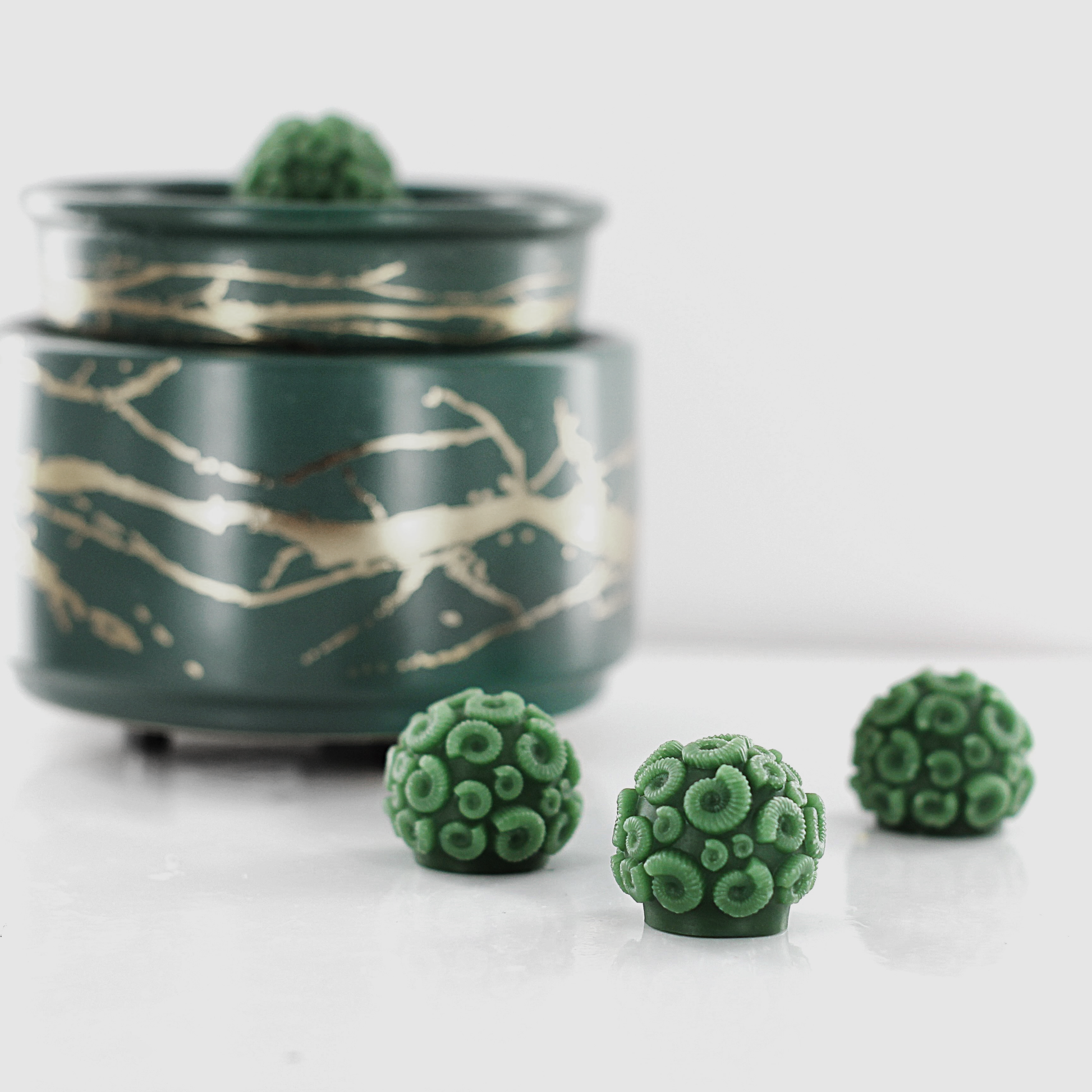 Green fragranced wax melts shaped like ammonites with intricate spiral details, displayed alongside a decorative wax warmer with a gold branch design.