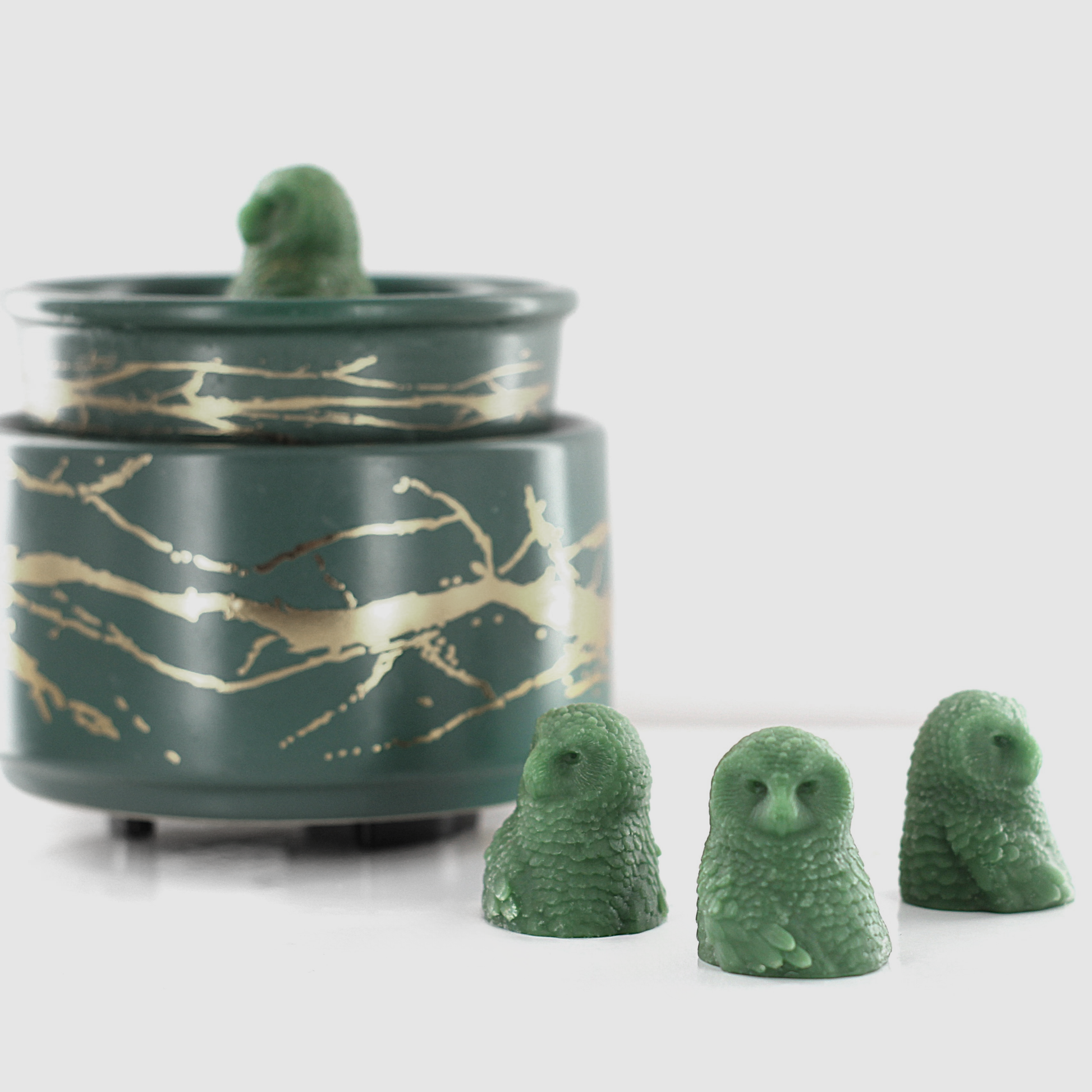 Green fragranced wax melts shaped like detailed owls, arranged near a decorative wax warmer with a gold branch design.