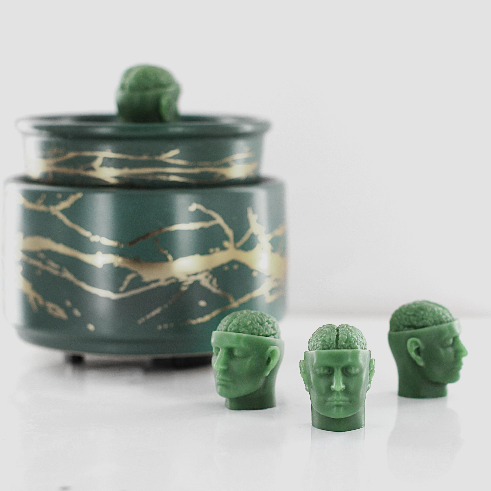 Green fragranced wax melts shaped like miniature human heads with exposed brains, displayed near a decorative wax warmer with a gold branch design.