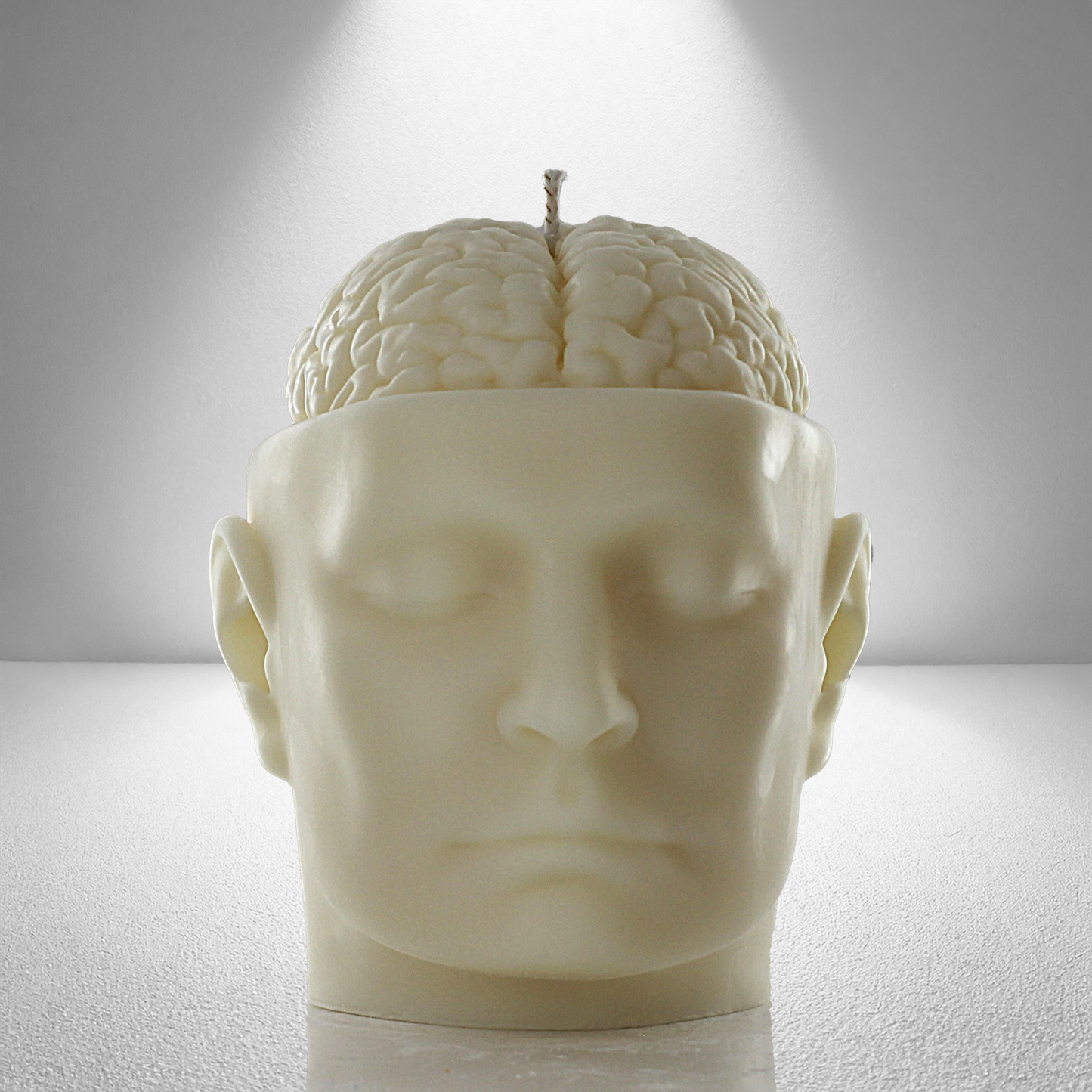 Front view of a soy candle shaped like a human head with an exposed brain design, featuring a wick on top. Cream-colored and detailed.