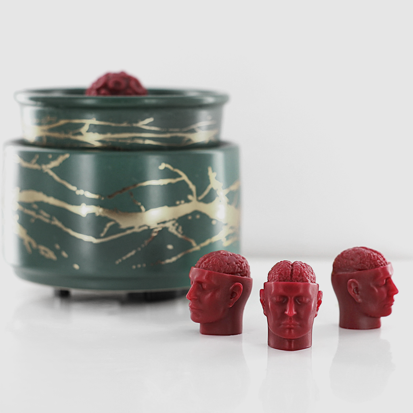 Red fragranced wax melts shaped like miniature human heads with exposed brains, displayed near a decorative wax warmer with a gold branch design.