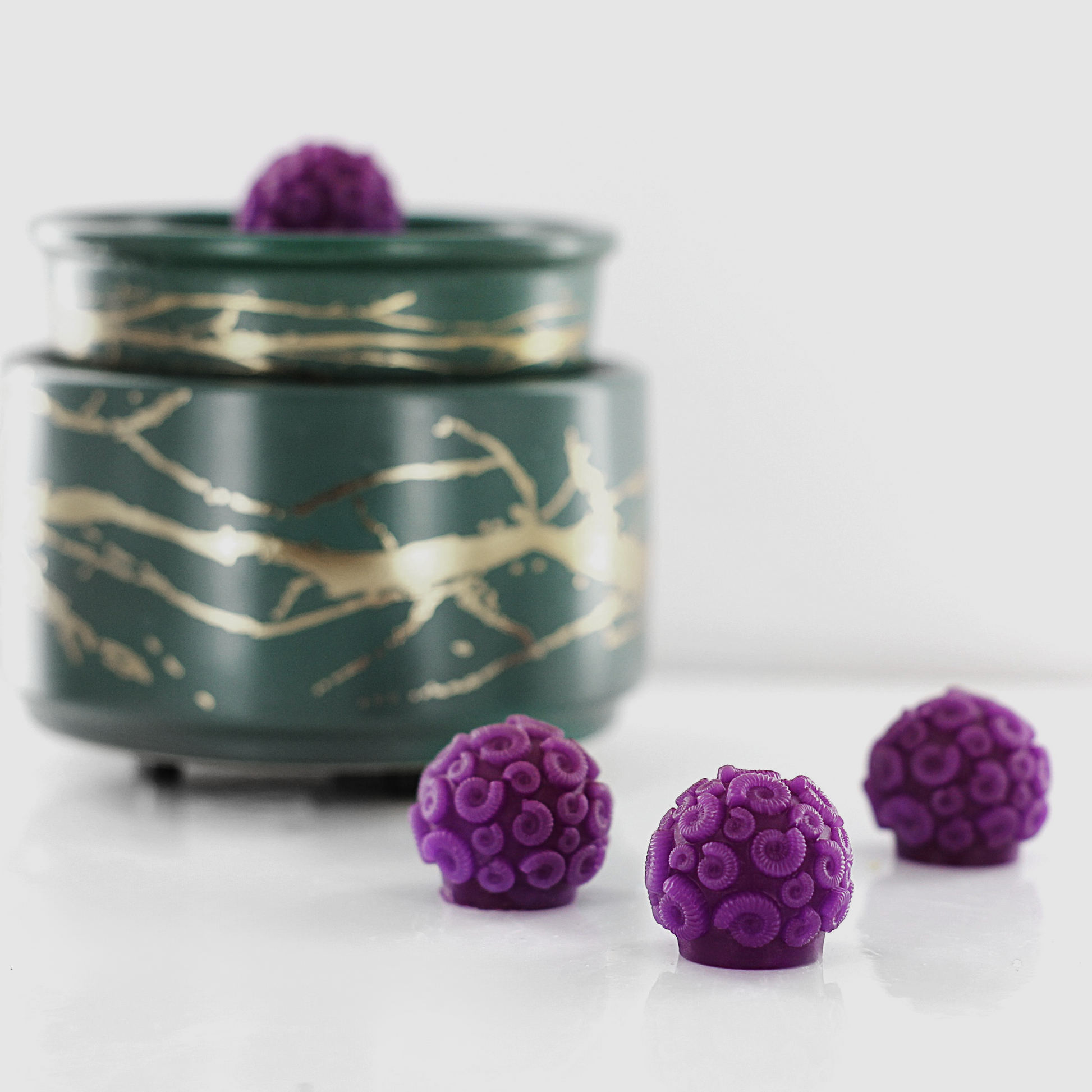 Purple fragranced wax melts shaped like ammonites with intricate spiral details, displayed alongside a decorative wax warmer with a gold branch design.