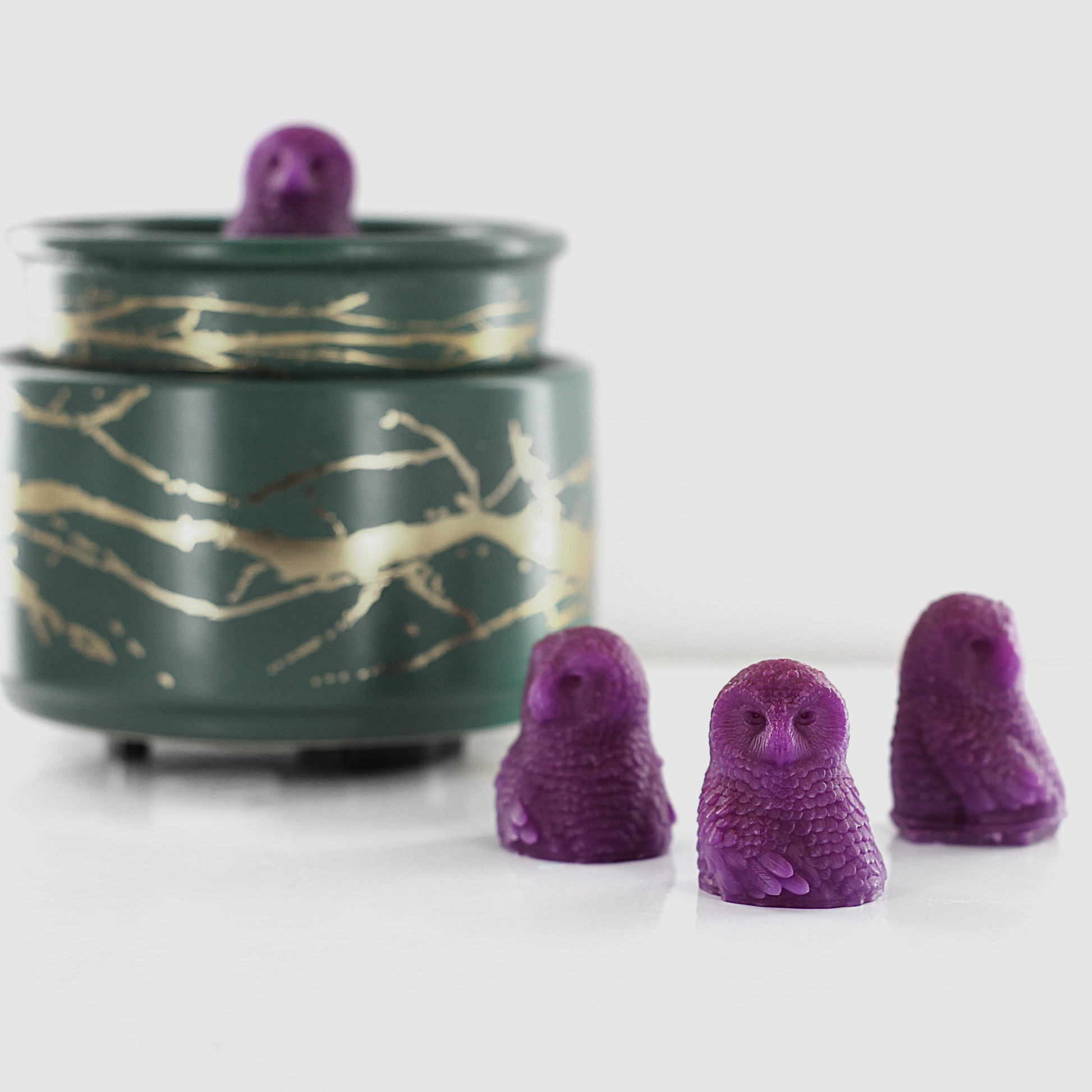 Violet fragranced wax melts shaped like detailed owls, arranged near a decorative wax warmer with a gold branch design.