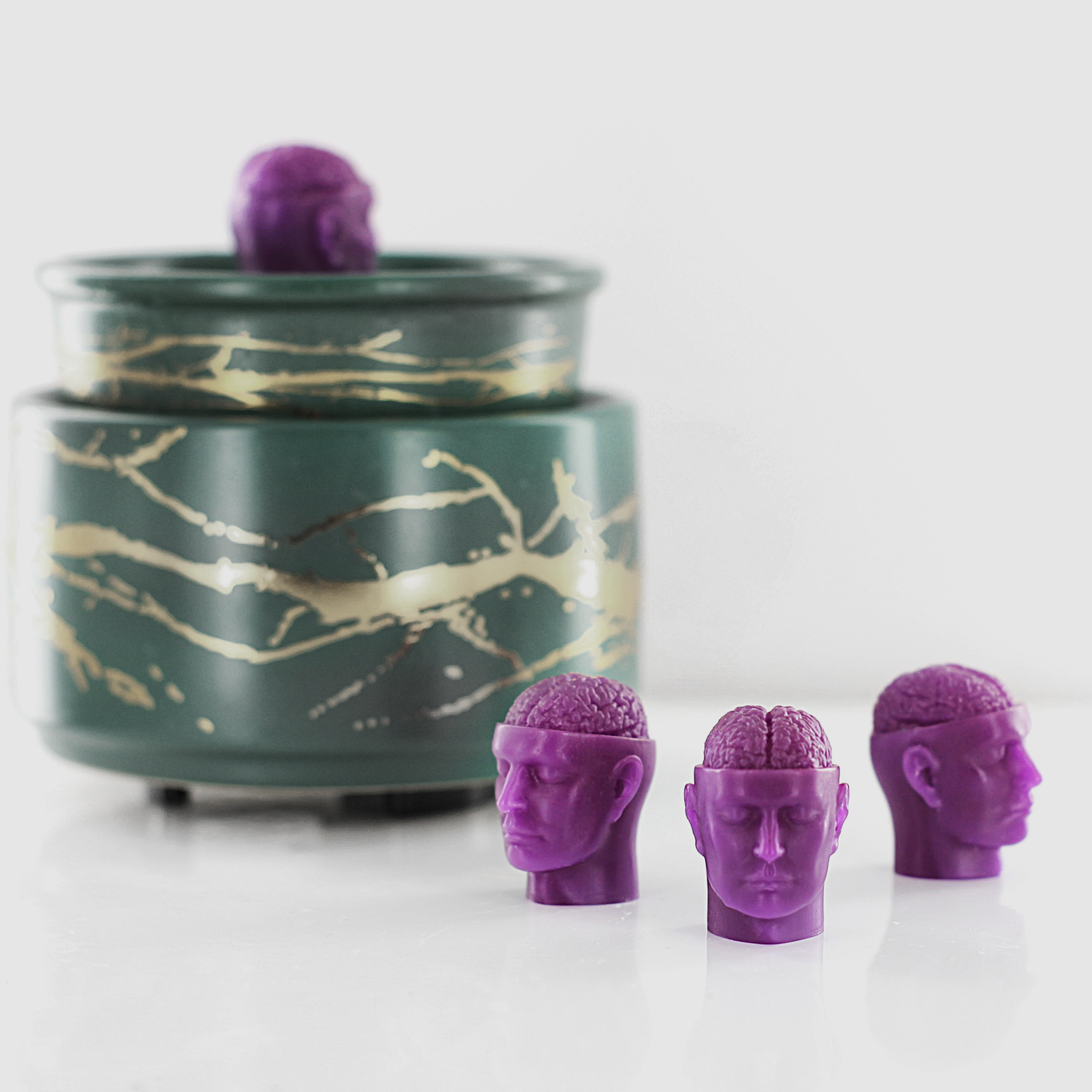 Purple fragranced wax melts shaped like miniature human heads with exposed brains, displayed near a decorative wax warmer with a gold branch design.